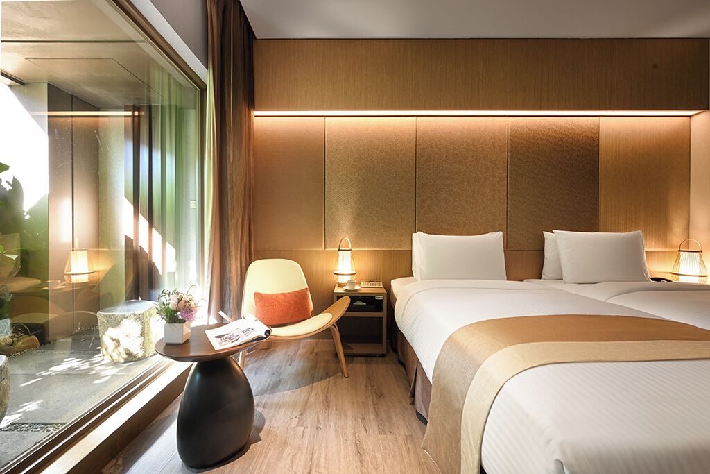 Una habitación de hotel con una cama grande, mesitas de noche, una silla y una pequeña mesa con una lámpara.
