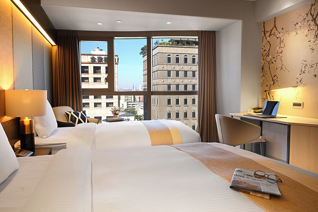 Une chambre d’hôtel avec deux lits, un bureau, une chaise, une lampe et une vue sur les bâtiments.