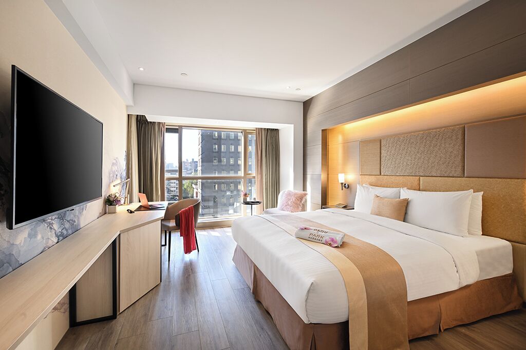 A hotel room with a large bed, a desk with a flat-screen TV, a chair, and a window with a city view.