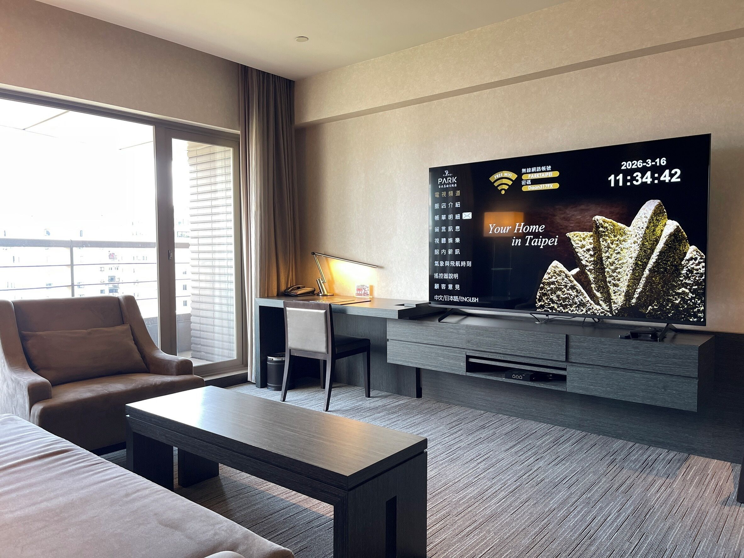 A modern hotel room with a large flat-screen TV, a desk, a comfortable sofa, and a large window.
