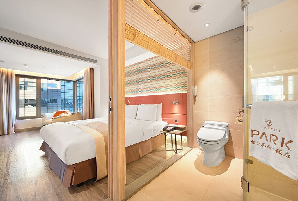 A hotel room with a large bed, a smaller bed, a bathroom with a toilet, and a view of the outside through a large window.