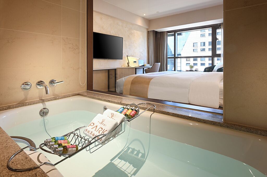 A hotel room with a large bathtub, a bed, a desk with a laptop, and a TV.