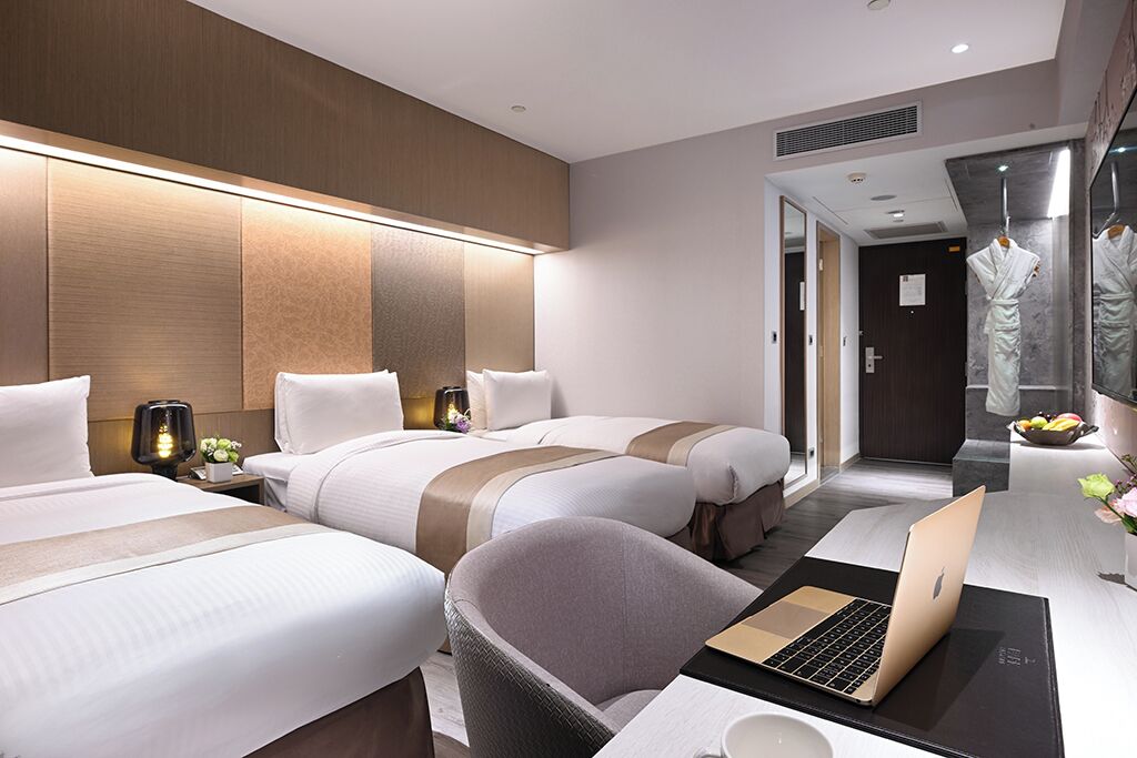 A hotel room with two beds, a desk with a laptop, a chair, and a bathroom with a mirror and robe.