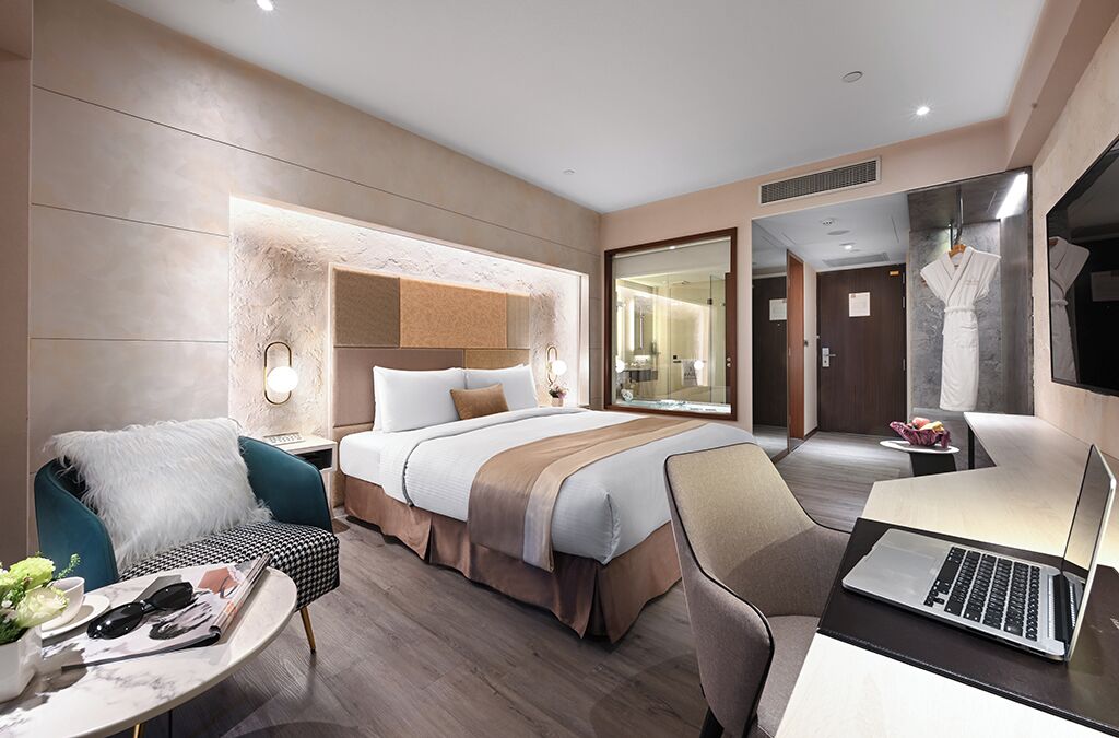 A modern hotel room with a large bed, a desk with a laptop, a chair, and a small table with a vase and remote control.