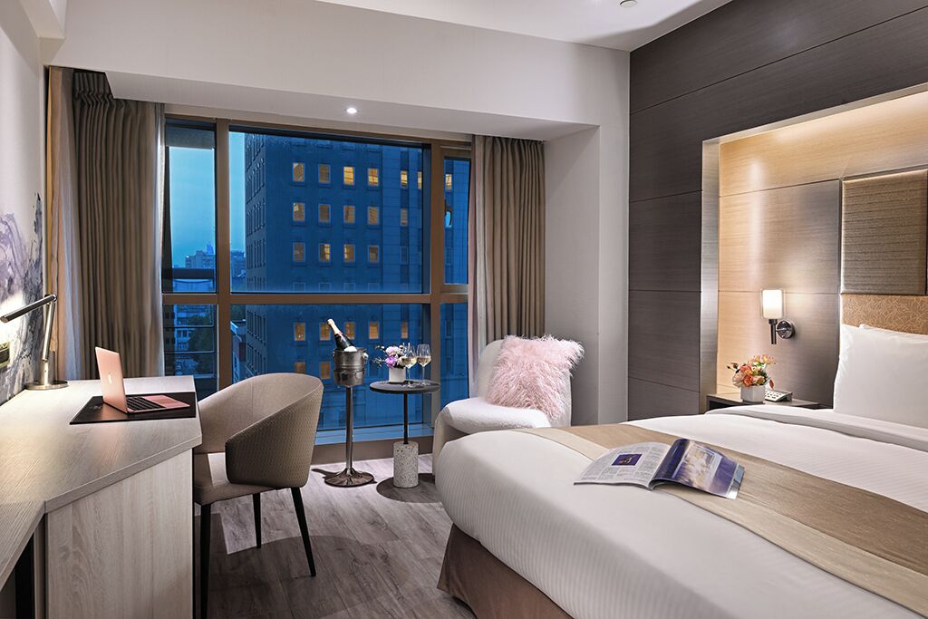 A modern hotel room with a large bed, a desk with a laptop, a small table with a drink and flowers, and a view of the city at night.