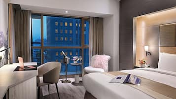 A modern hotel room with a large bed, a desk with a laptop, a small table with a drink and flowers, and a view of the city at night.