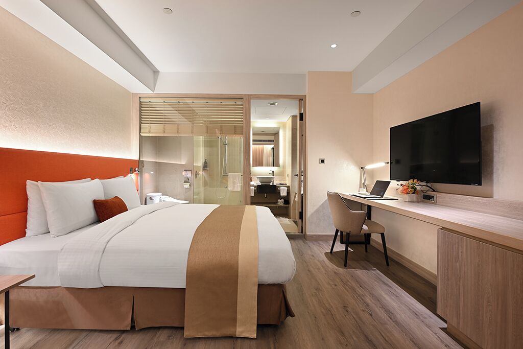 A hotel room with a bed, a desk, a TV, and a bathroom visible through a glass door.