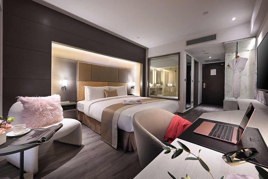 Habitación de hotel moderna con una cama grande, un escritorio con una laptop, una silla y una pequeña mesa con una taza y flores.
