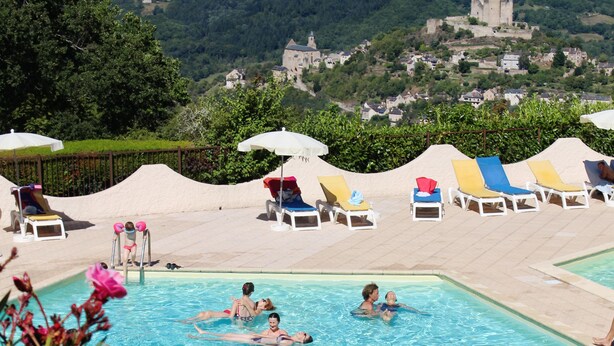 A swimming pool with people swimming, surrounded by lounge chairs and a view of a castle on a hill.