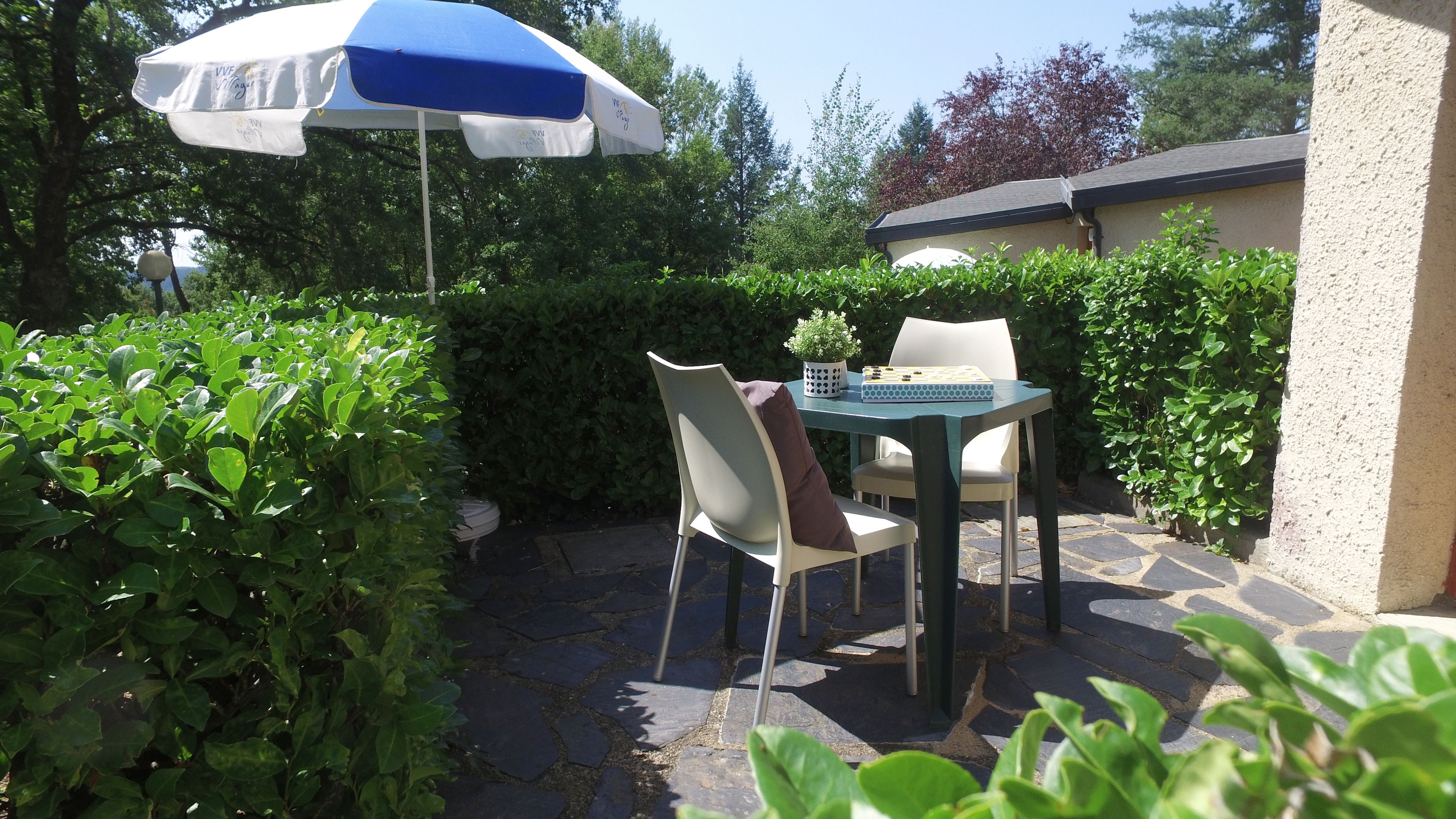 Un patio exterior con una mesa y sillas, un paraguas azul y blanco, y abundante follaje.