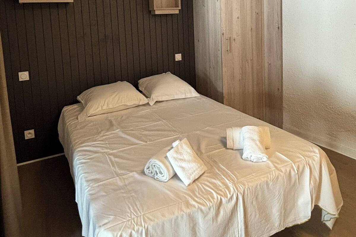 A single bed with white linens and two pillows, a wooden wardrobe, and two small wooden cabinets against a dark wall.