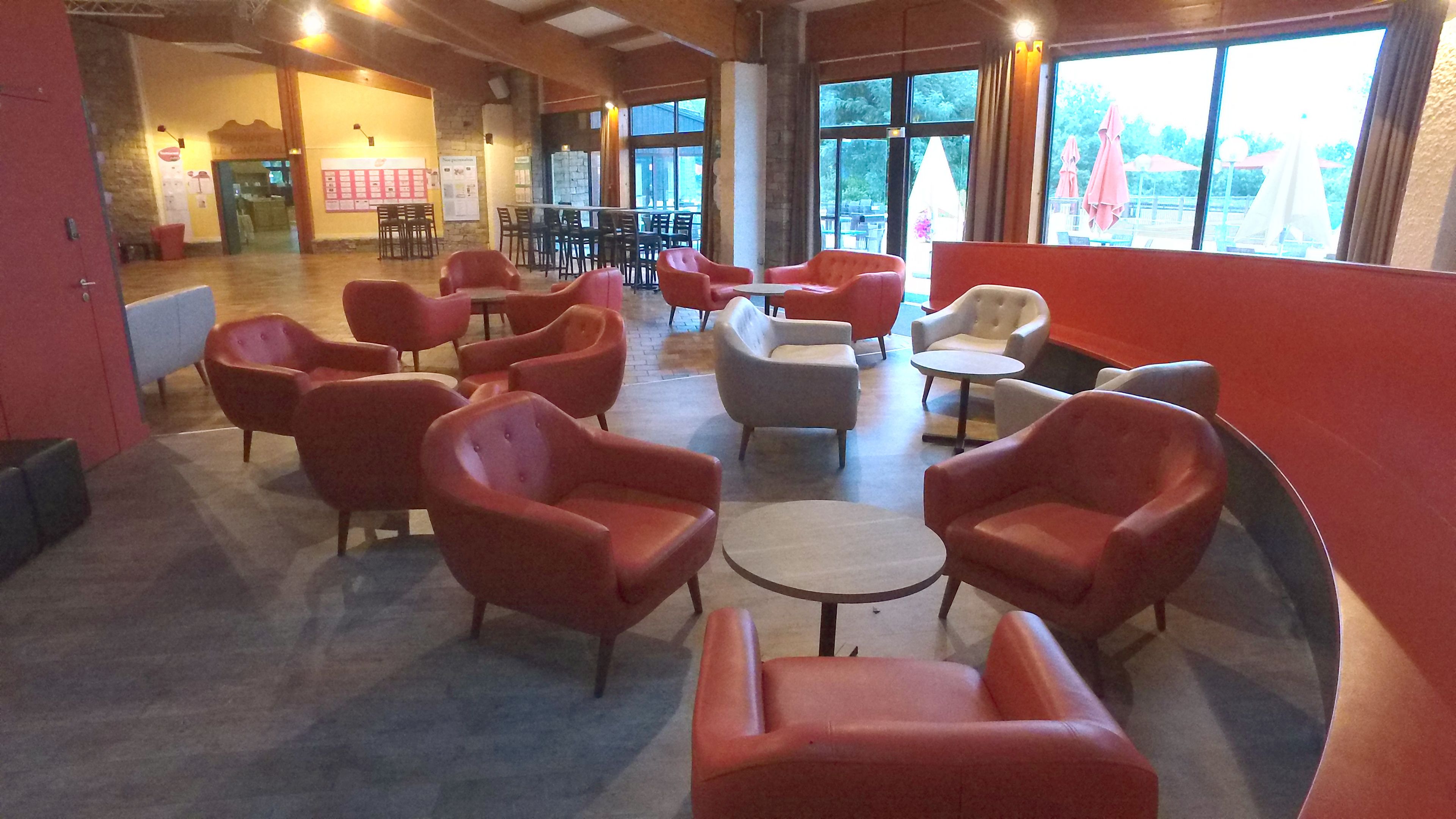 Un vaste espace intérieur aménagé avec différents types de sièges, notamment des chaises et des tables rouges et grises. De grandes fenêtres laissent entrer la lumière naturelle et offrent une vue sur l’extérieur.