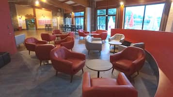 Un vaste espace intérieur aménagé avec différents types de sièges, notamment des chaises et des tables rouges et grises. De grandes fenêtres laissent entrer la lumière naturelle et offrent une vue sur l’extérieur.