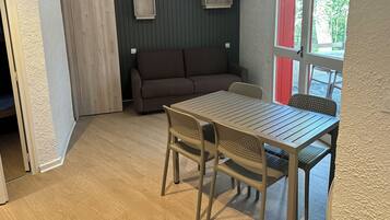 Una habitación con piso de madera, un sofá marrón, una mesa con sillas y dos estantes en la pared.