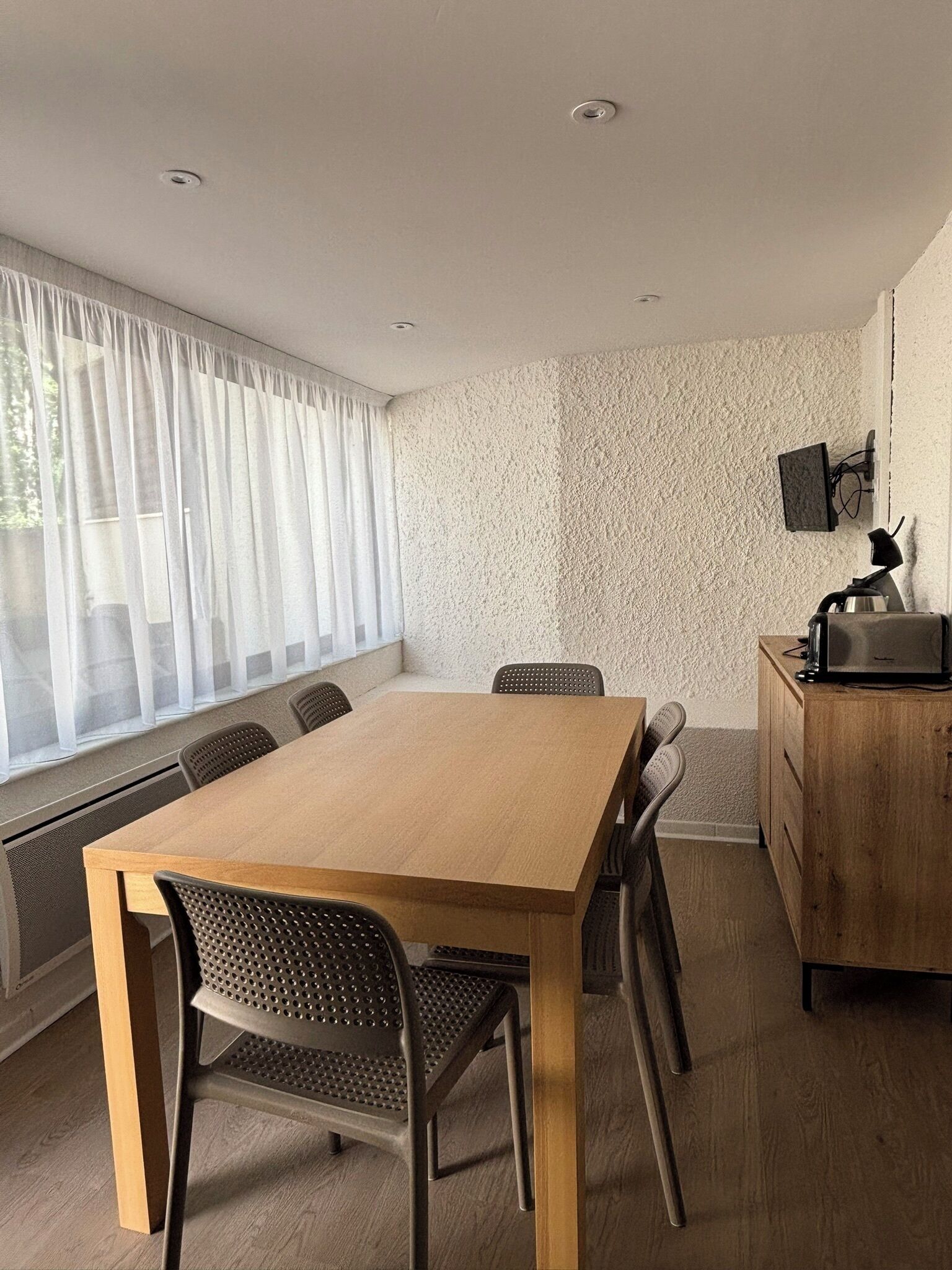 Un espace repas moderne avec une table et des chaises en bois, un téléviseur fixé au mur et une fenêtre agrémentée de rideaux légers.