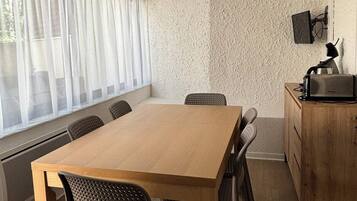Un espace repas moderne avec une table et des chaises en bois, un téléviseur fixé au mur et une fenêtre agrémentée de rideaux légers.