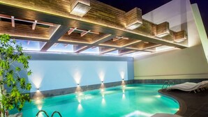 An indoor swimming pool with a modern design, featuring a clear blue water surface, a sunken seating area, and a ceiling with wooden beams and integrated lighting. - Capitol Hotel (Dubai)