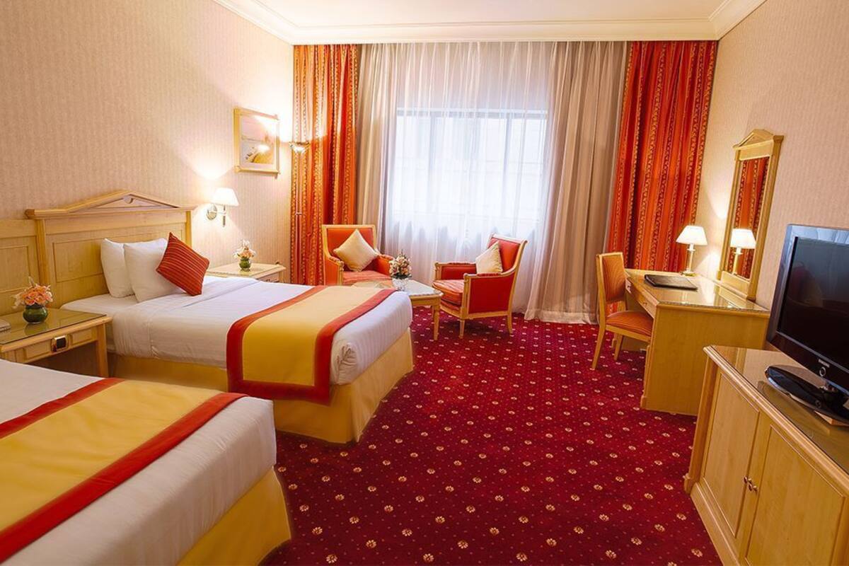 Habitación de hotel con dos camas, un escritorio, una silla, un televisor y un ventanal grande con cortinas.