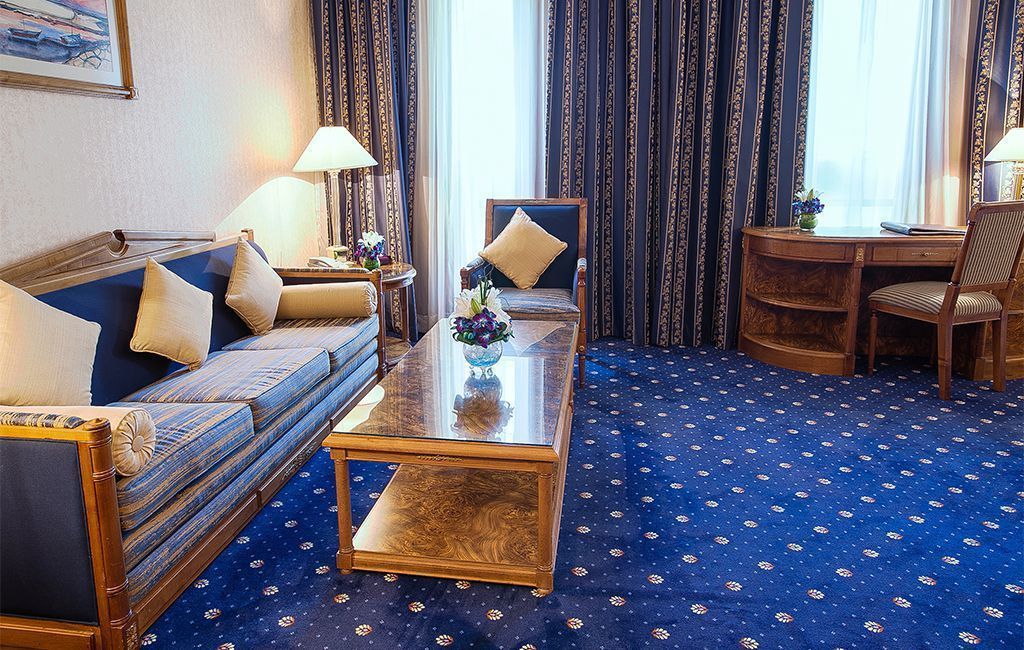 Hotellihuone, jossa on sinikuvioinen matto, tyynyillä varustettu sohva, puinen sohvapöytä, työpöytä tuolilla sekä kuvioidut verhot.