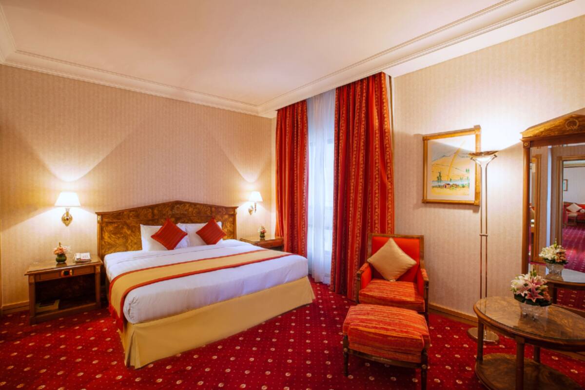 Una habitación de hotel con una cama grande, cortinas rojas, un cabecero de madera, una mesita con flores y un espejo en la pared.