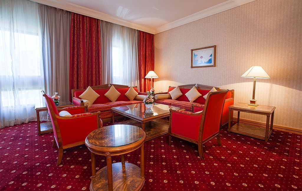 A hotel room with a red carpet, a sofa set, two lamps, and a painting on the wall.
