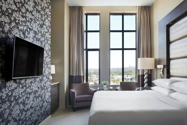 Habitación de hotel con una cama grande, un televisor montado en la pared, un escritorio con lámpara, dos sillas y vistas a la ciudad a través de amplios ventanales.
