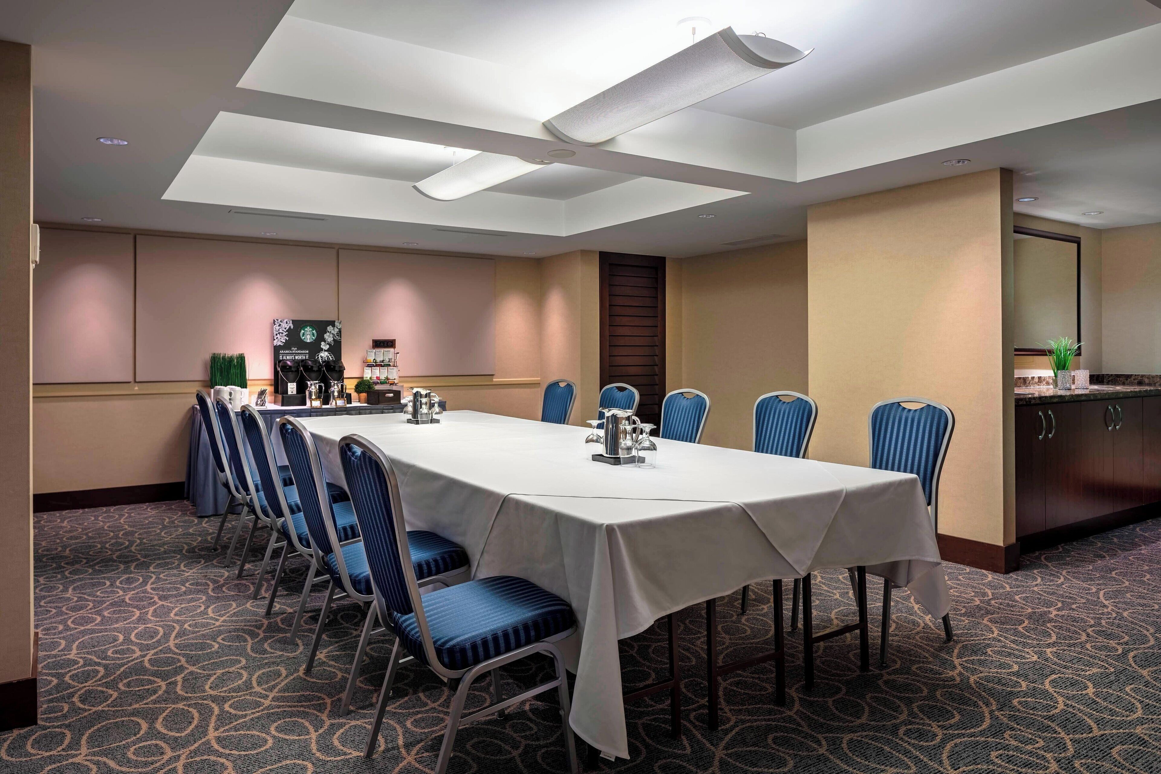 A conference room with a long table, blue chairs, a white tablecloth, and a kitchen area in the background.