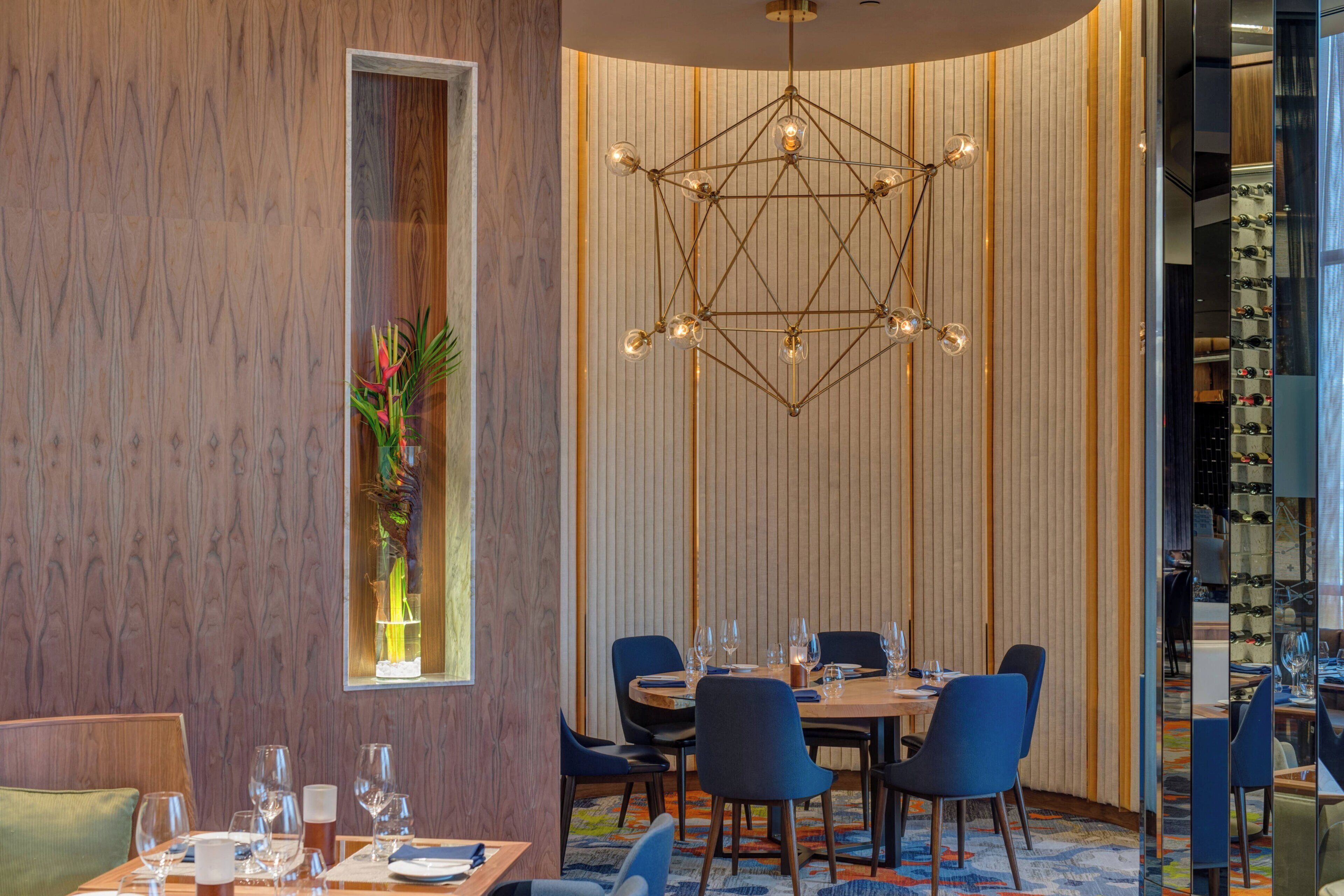 Un espace de salle à manger moderne, agrémenté d’un lustre aux motifs géométriques, de murs recouverts de panneaux en bois et offrant une vue sur un meuble à vin.