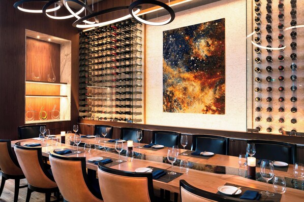 Un restaurante con una larga mesa de comedor, varias estanterías para vino y un cuadro abstracto grande en la pared.