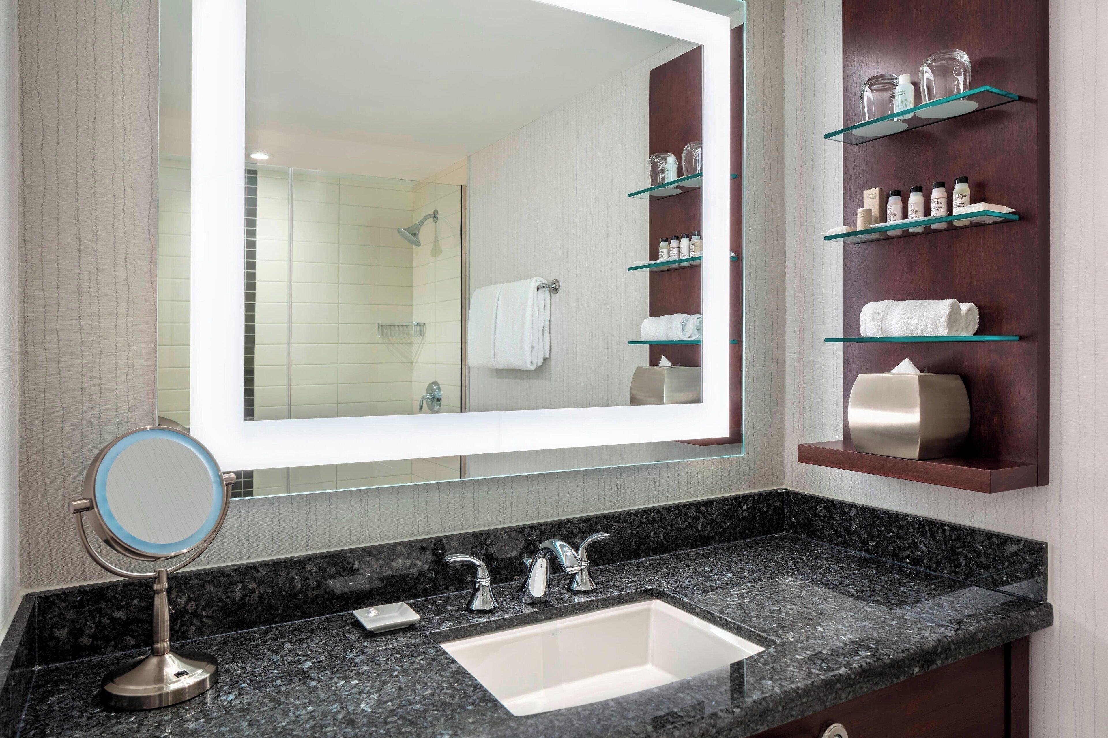 A modern bathroom with a large mirror, a granite countertop, a sink, and shelves with toiletries.