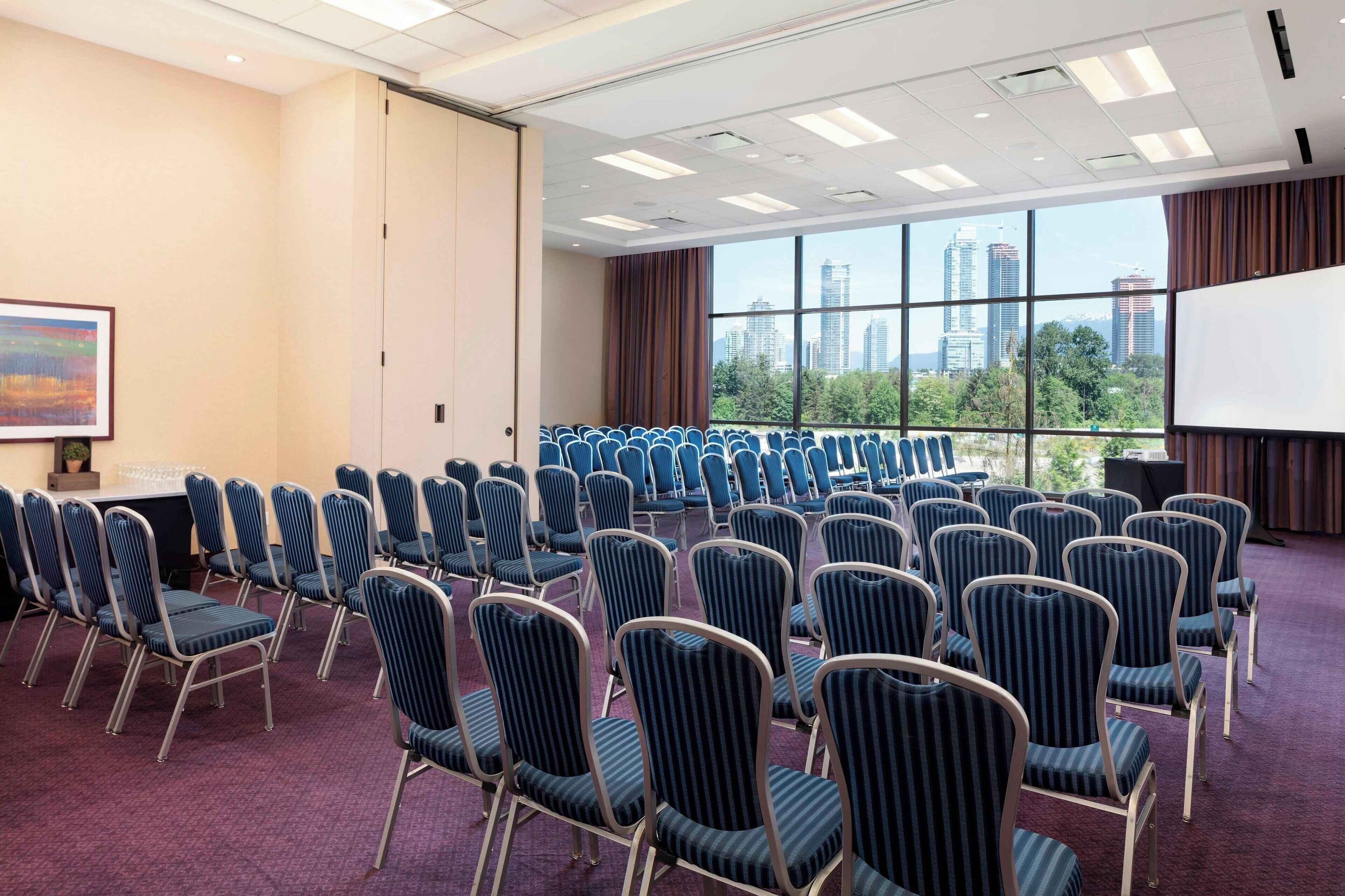 A conference room with rows of chairs, a projector screen, and a large window with a city view.