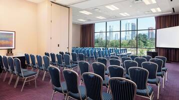 A conference room with rows of chairs, a projector screen, and a large window with a city view.