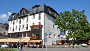 A hotel with a balcony adorned with flowers, a sign indicating a restaurant, and a street with people dining outside.