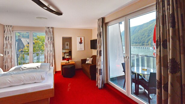 A hotel room with a bed, a desk, a chair, a balcony with a table and chairs, and a view of a bridge and hills.