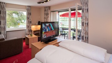 Habitación de hotel con cama, televisión, pequeña mesa con lámpara, silla y vista al exterior a través de una puerta corrediza.