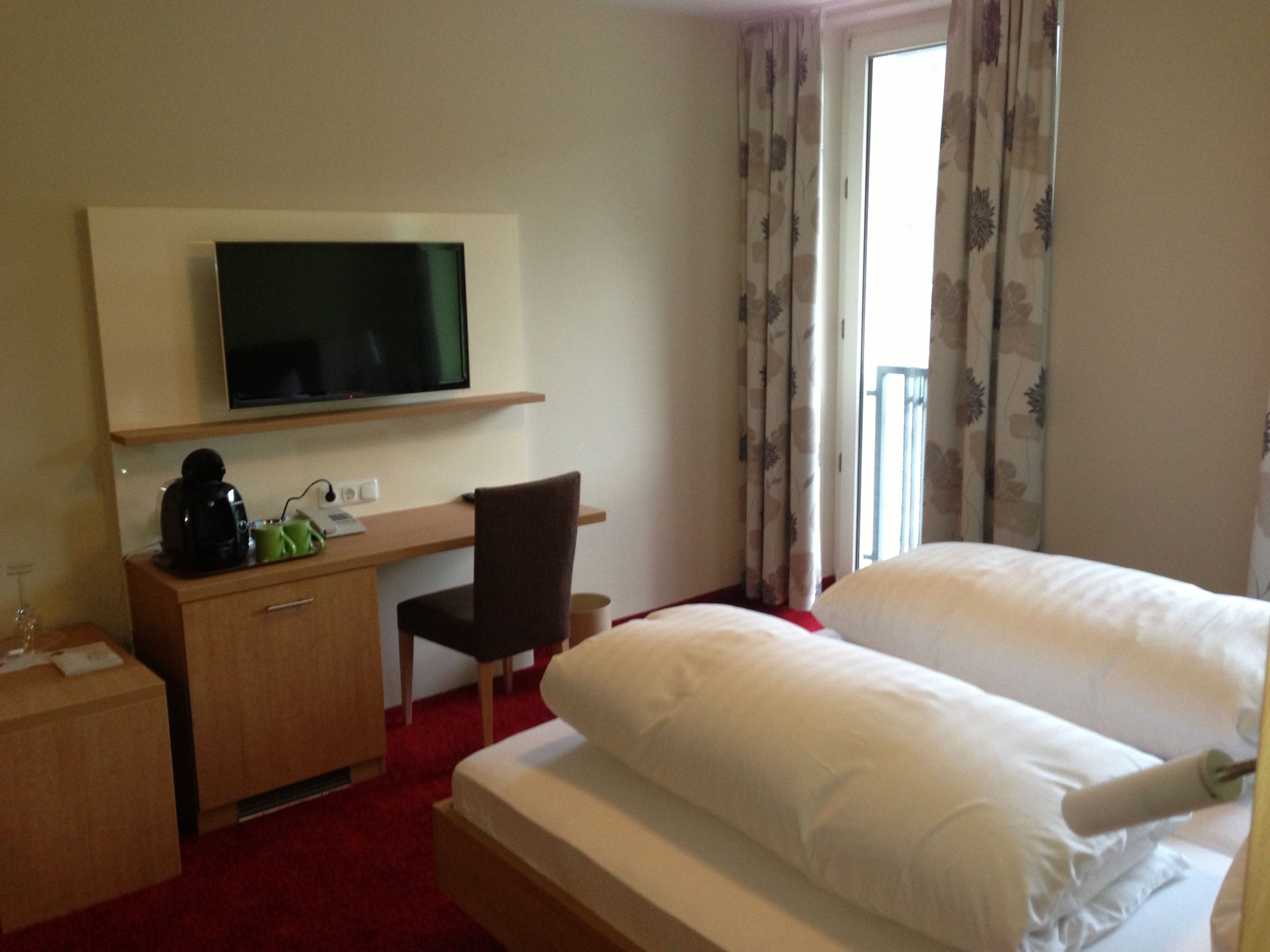 A hotel room with a bed, a desk with a chair, a television, and a window with curtains.