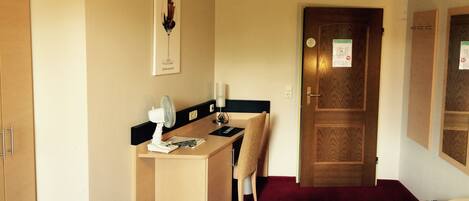 A hotel room with a bed, a desk, a chair, a fan, and a door with a sign.