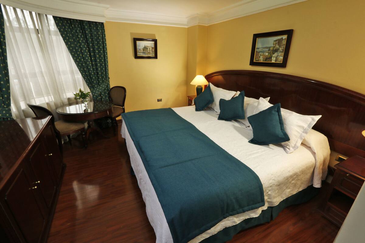 Habitación de hotel con una cama grande, un escritorio y una silla. Hay dos cuadros en las paredes y una lámpara en la mesita de noche.