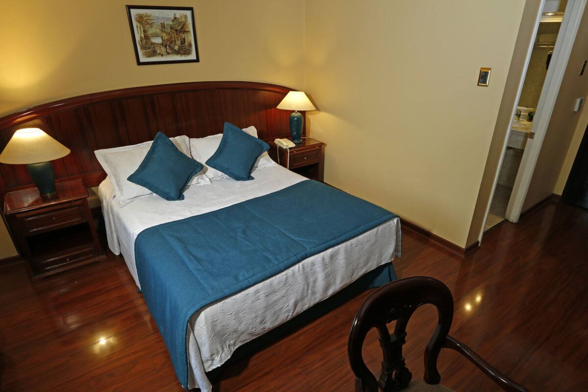 Una cama bien hecha con ropa de cama azul, dos mesitas de noche con lámparas y una silla de madera en una habitación de hotel.