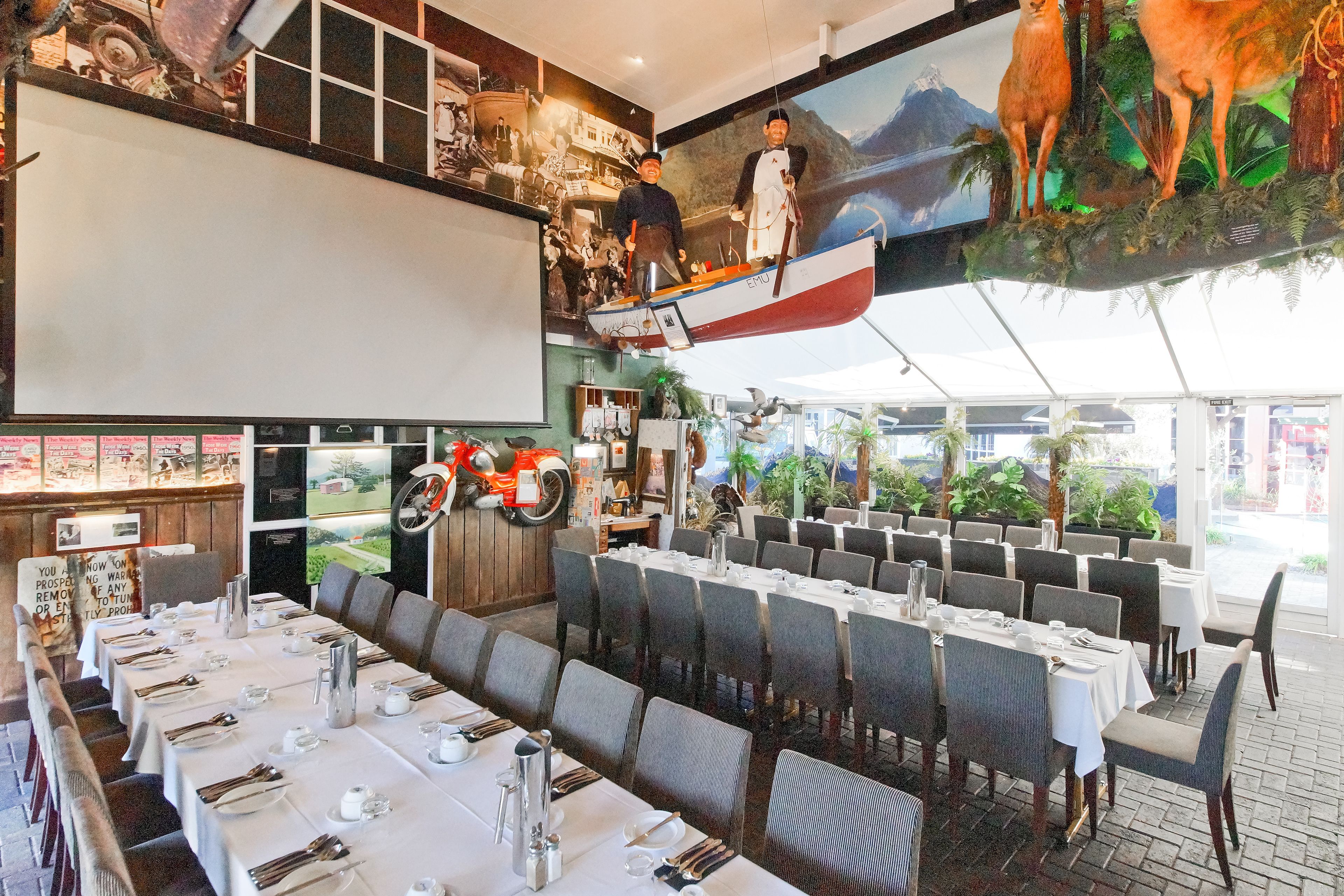 A restaurant with long tables set for dining, a motorcycle on the wall, and a large mural of a man on a boat.