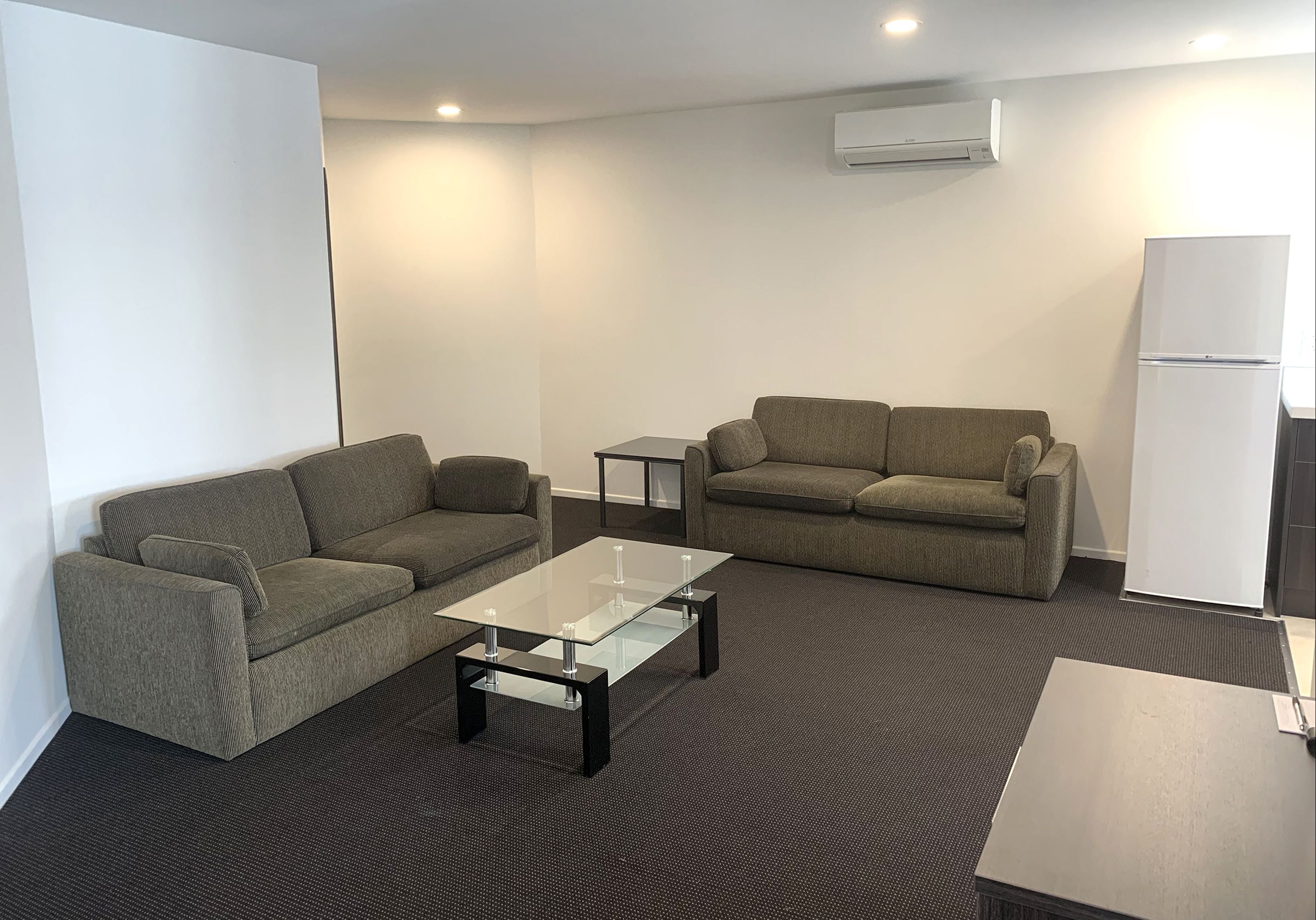 Una sala de estar moderna con dos sofás, una mesa de centro de vidrio, una mesita auxiliar y un refrigerador.
