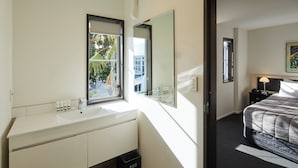 Ett modernt hotellrum med en vit sminkbord, en stor spegel, en säng med ett grått täcke och ett sängbord med en lampa.