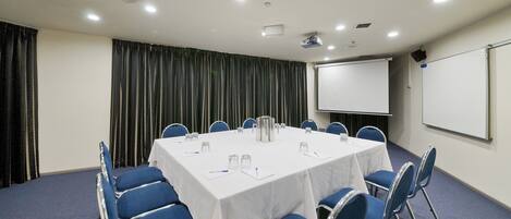 A conference room with a round table, blue chairs, a white tablecloth, a projector screen, and a whiteboard.