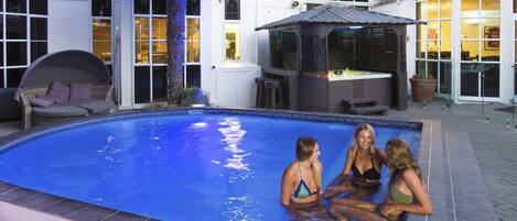 A swimming pool with three people sitting on the edge, a hot tub, and a covered seating area.