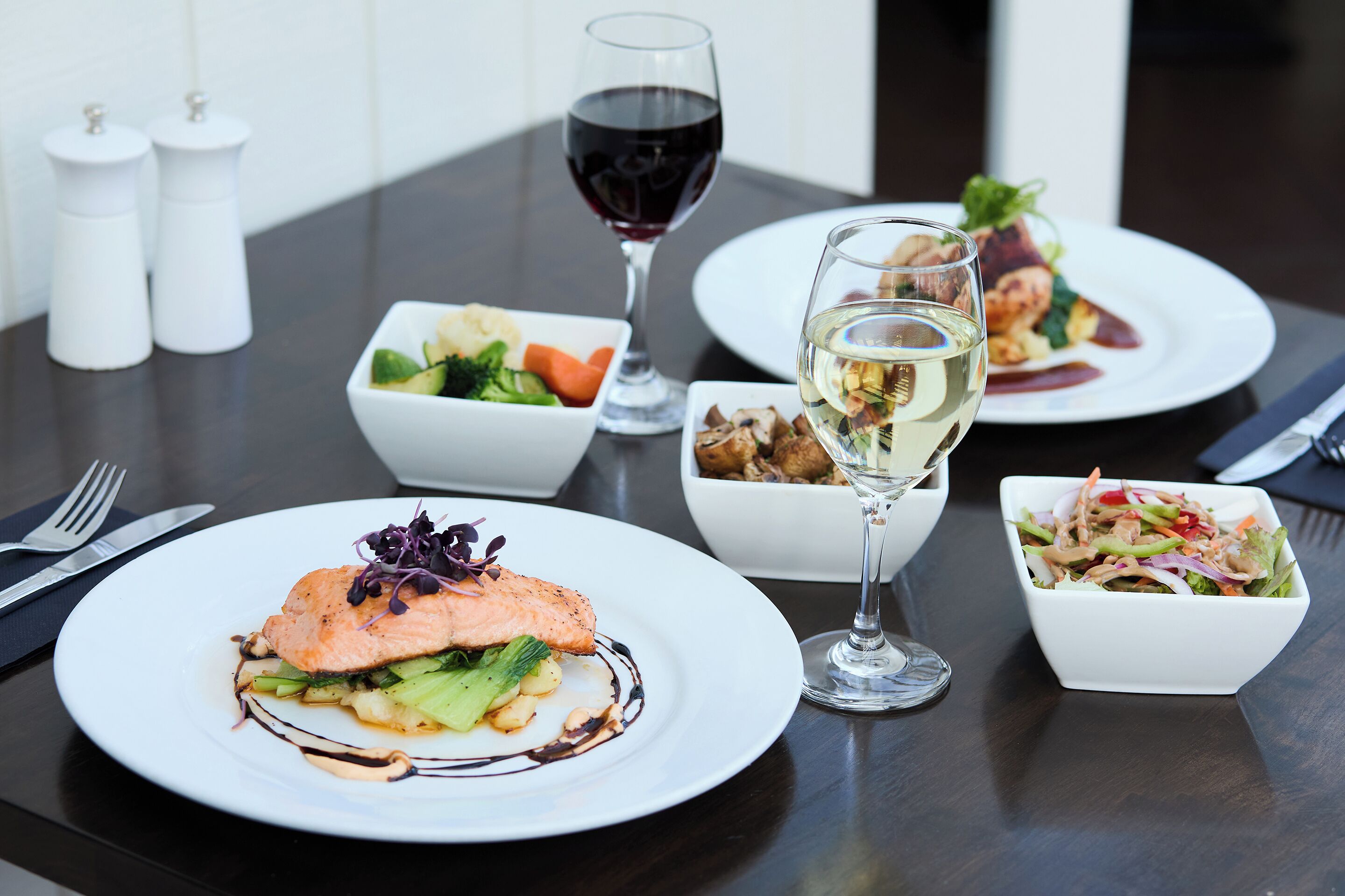 A dining table with three plates of food, two glasses of wine, and salt and pepper shakers.