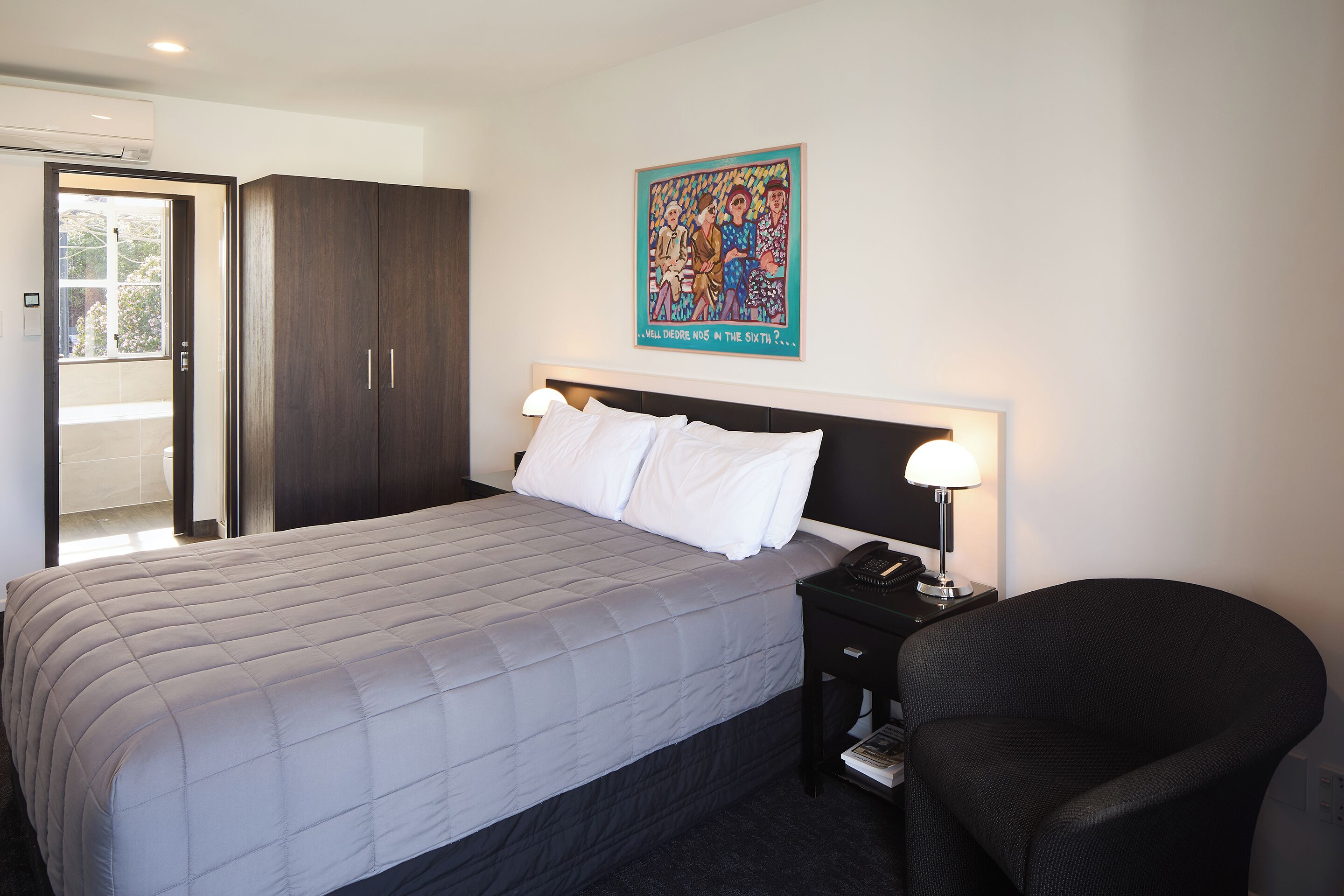 Habitación de hotel con una cama, una mesita de noche, una silla, una lámpara fijada a la pared y una puerta que da a un balcón.