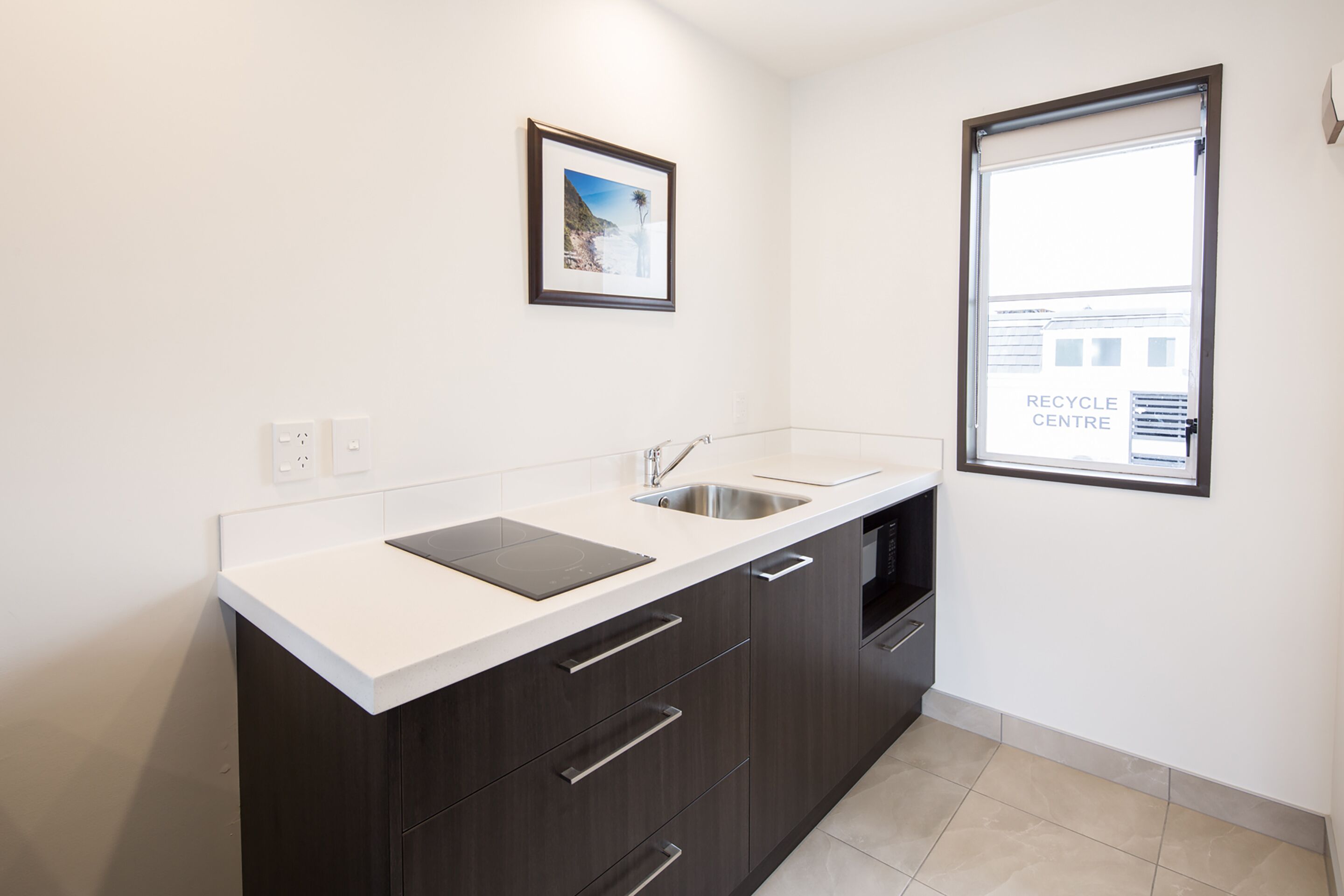 A modern kitchen with dark cabinetry, a white countertop, a stainless steel sink, and a framed picture on the wall.