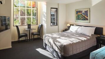 A hotel room with a large bed, a desk with a chair, a small table, and a nightstand with a lamp.