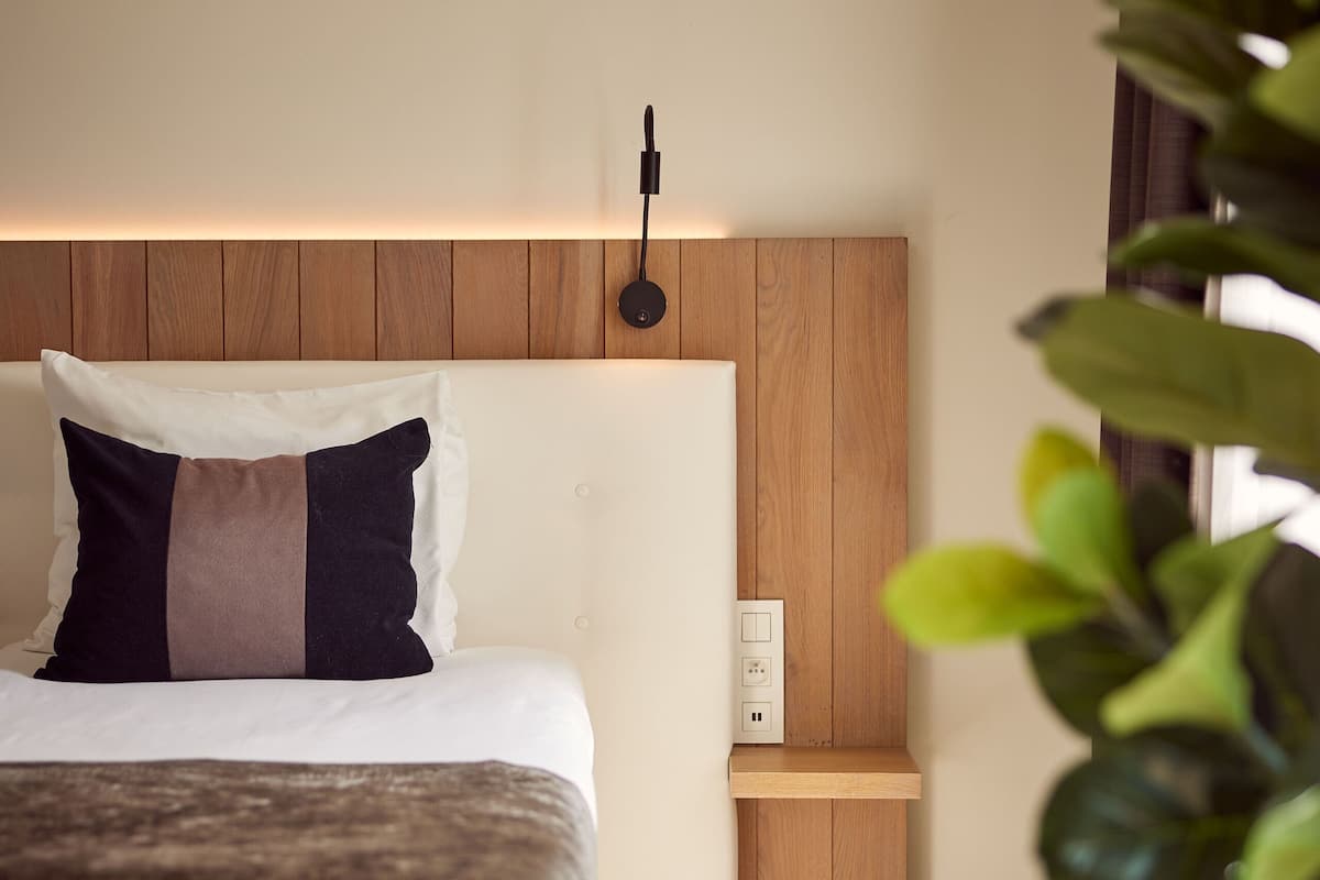 Une chambre d’hôtel moderne avec une tête de lit en bois, un lit recouvert d’une couverture marron, une table de chevet et une lampe fixée au mur.