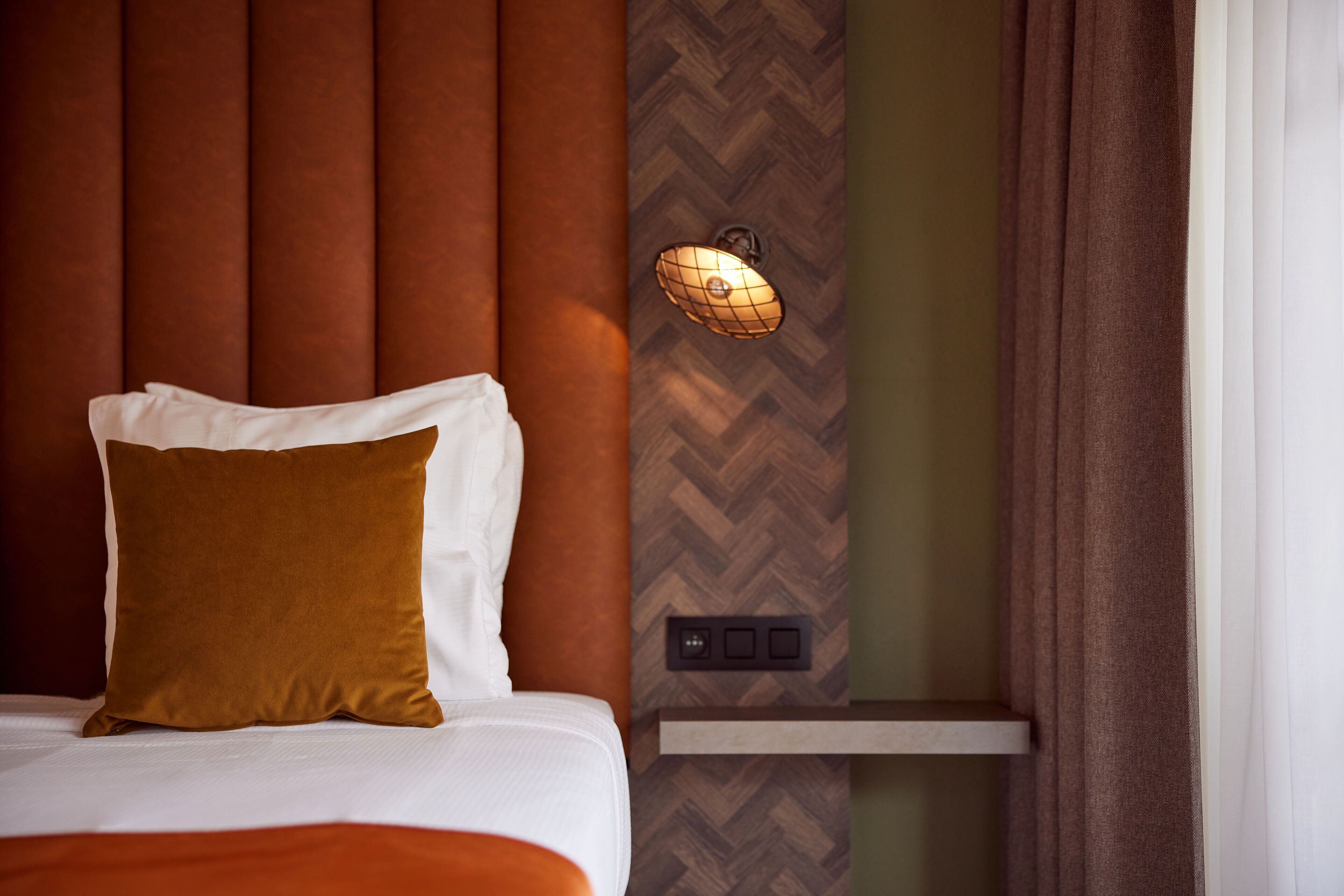 A hotel room with a bed, a brown pillow, a wall-mounted light fixture, and a control panel.